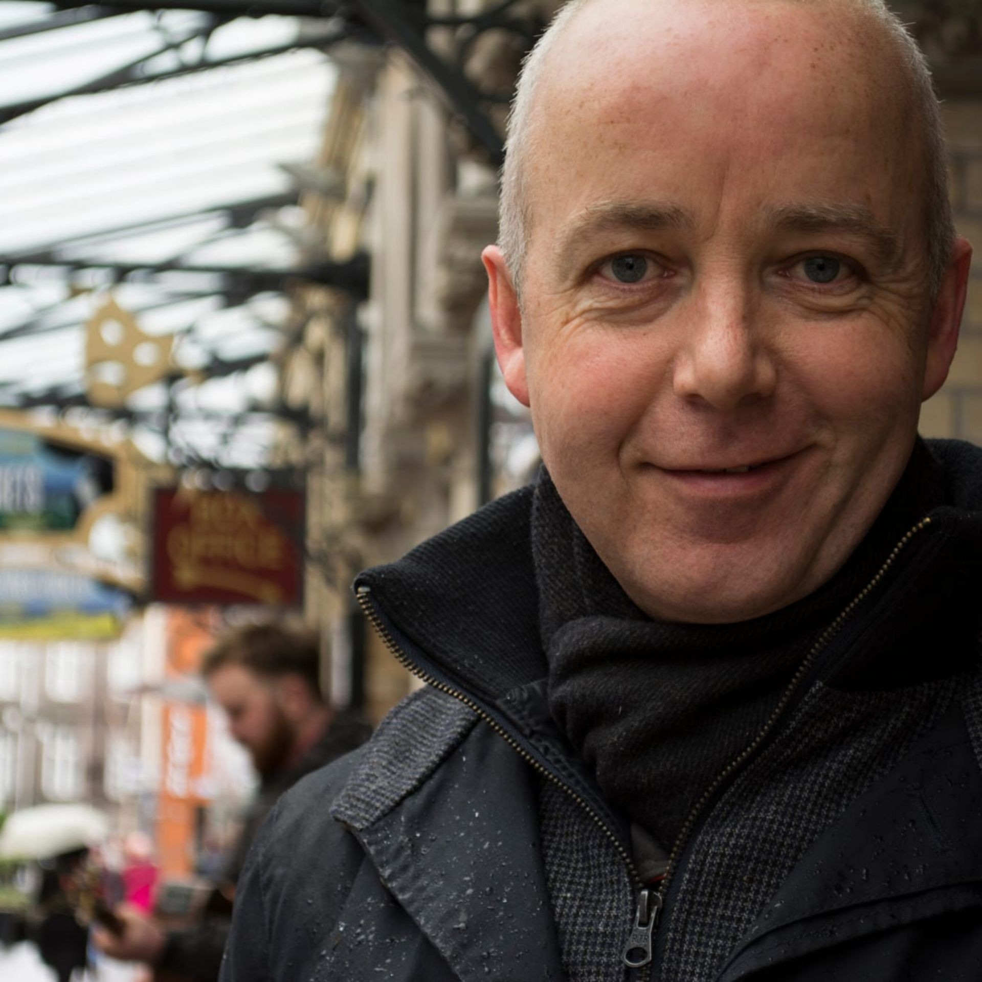 Man smiling outdoors under a metal canopy with blurred background of signs and another person.